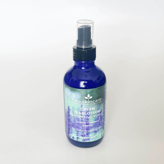 A 4-ounce glass mist spray bottle of Fate Skincare's Green Teablossom Hydrating Tonic in a gradient of green, turquoise, blue, and purple hues. The bottle rests elegantly in a field of lavender reeds, blending harmoniously with the vibrant colors of the background. The label is prominently displayed, highlighting its organic and cruelty-free ingredients, evoking a sense of natural freshness and serenity.