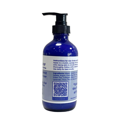 A closer look at the side of the blue glass bottle. The label includes usage instructions: "Shake well before use. Apply to a loofah, sponge, or hands, then massage onto damp skin in circular motions. Rinse thoroughly. Follow with a moisturizer for best results." The ingredient list includes water, coconut, shea butter, jojoba, argan, aloe vera, green tea, collagen, bakuchiol, and essential oils such as lavender and rose. A QR code is present for additional details.