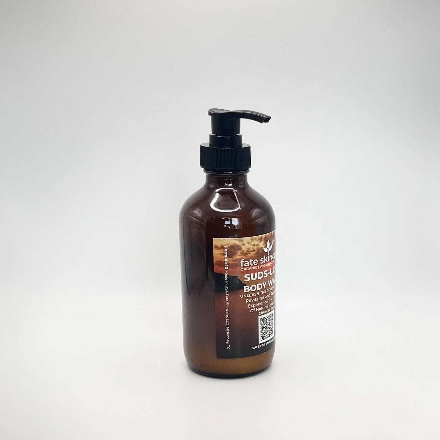 Fate Skincare's Suds-less Body Wash in a 8 ounce glass amber bottle equipped with a black pump dispenser. The label prominently displays the product's name in white extra-bold letters against a background of a golden sky.