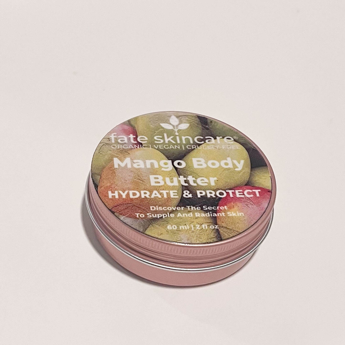 Round rose gold tin of Fate Skincare's 2 oz Mango Body Butter on a plain white background. The label displays mangoes, the product name, quantity, and Fate Skincare's logo.