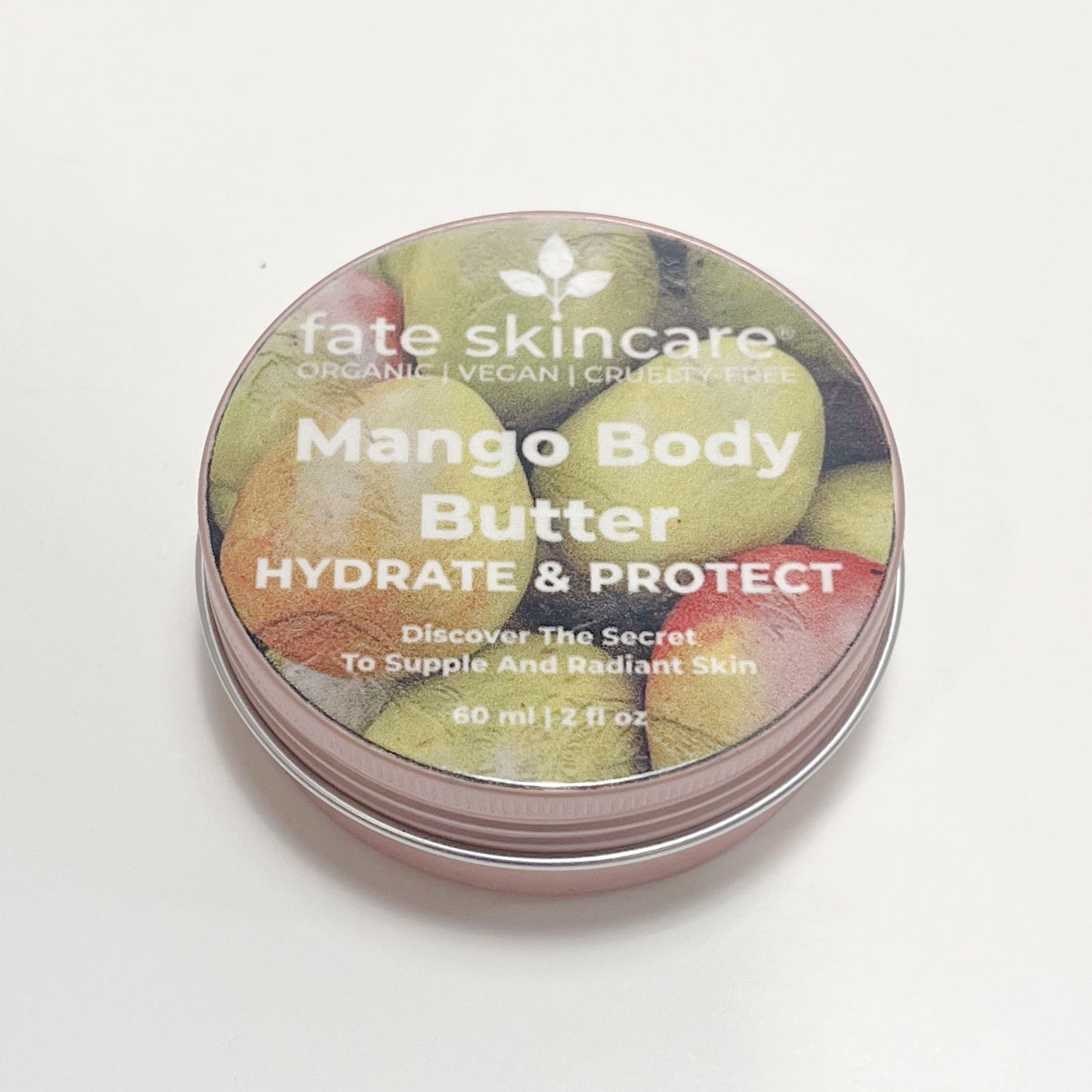 A round, rose-colored aluminum tin of Fate Skincare Mango Body Butter. The lid features an image of ripe mangoes in the background with white text stating "Hydrate & Protect." The label promotes its benefits for supple and radiant skin, and the container holds 60 ml (2 fl oz) of product.